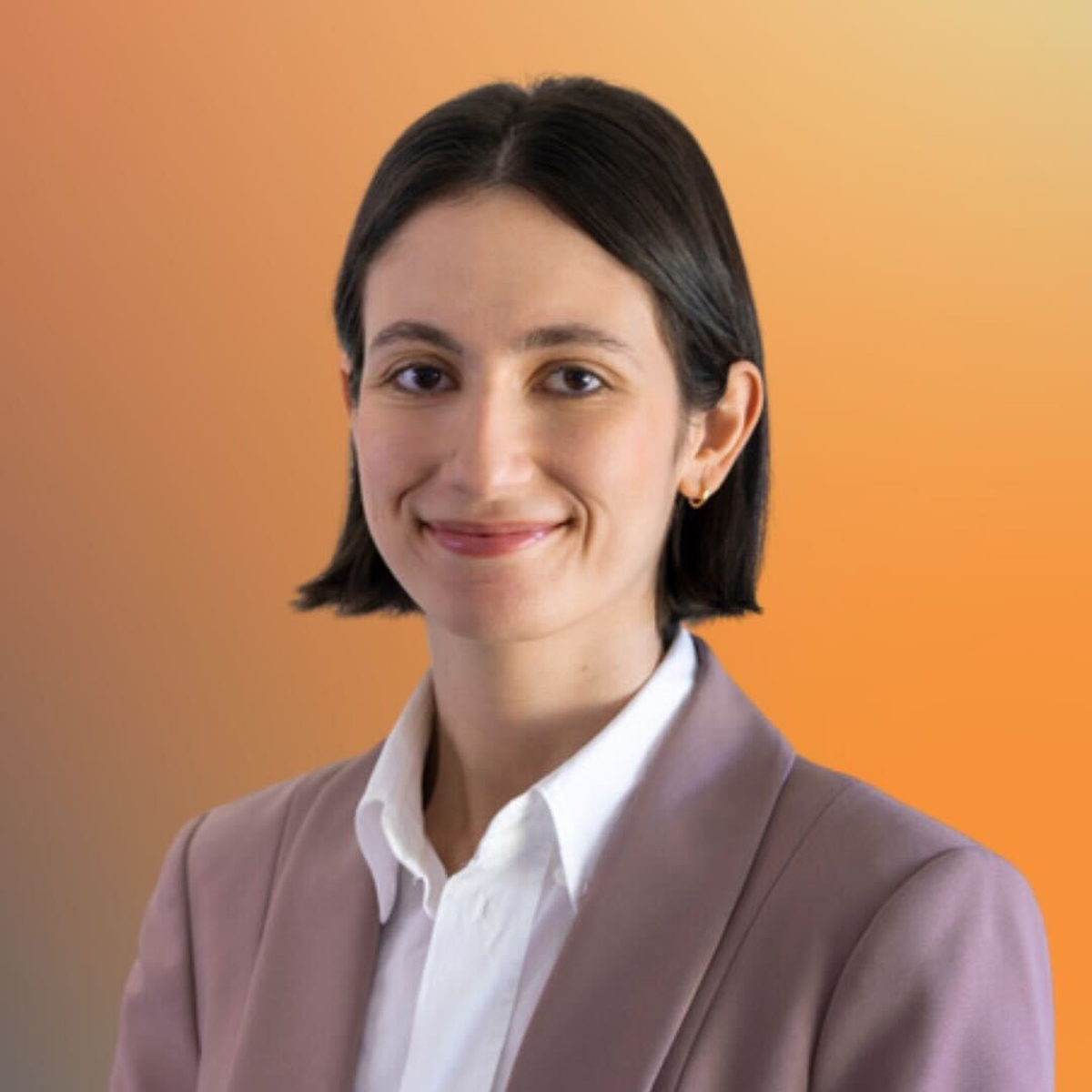 Smiling person with short dark hair in a lavender blazer and white shirt, set against a soft orange gradient background.