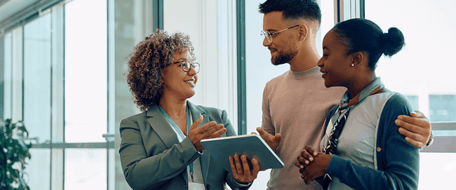 Three people smiling and talking in a bright office, with one holding a tablet. Large windows in the background.