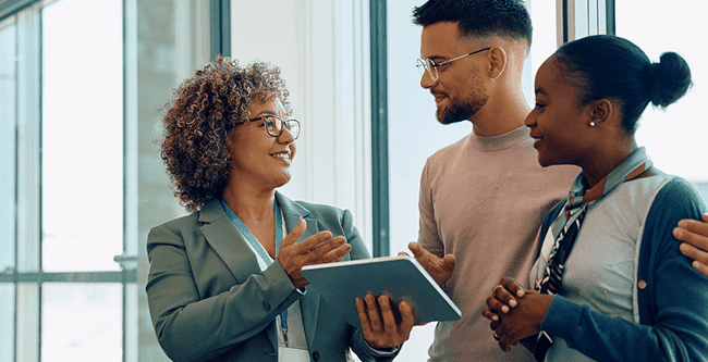 Three people smiling and talking in a bright office, with one holding a tablet. Large windows in the background.