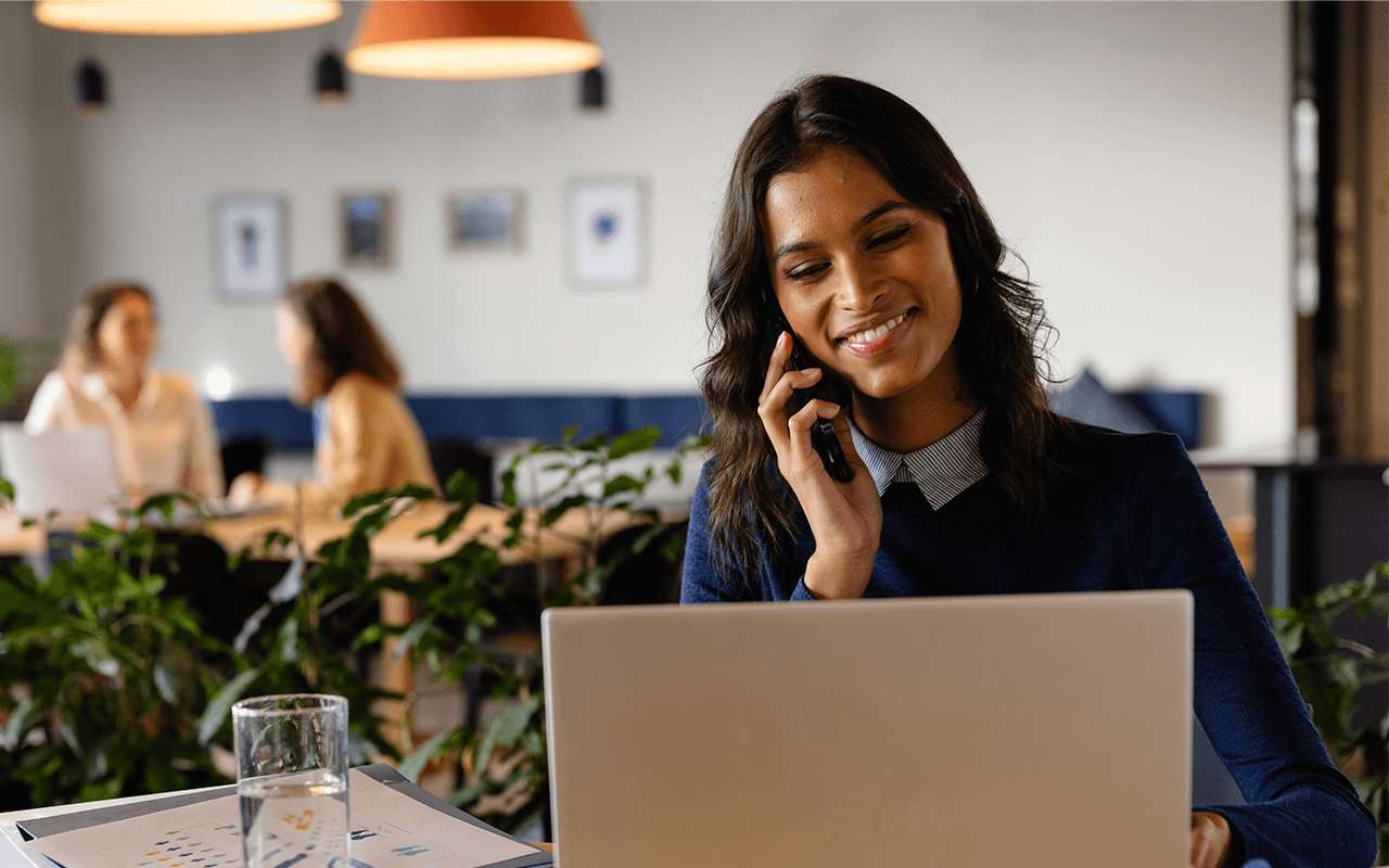 Woman smiling while talking on phone, seated at desk with laptop in a cozy office space. Two people converse in the background. Plants and decor visible.
