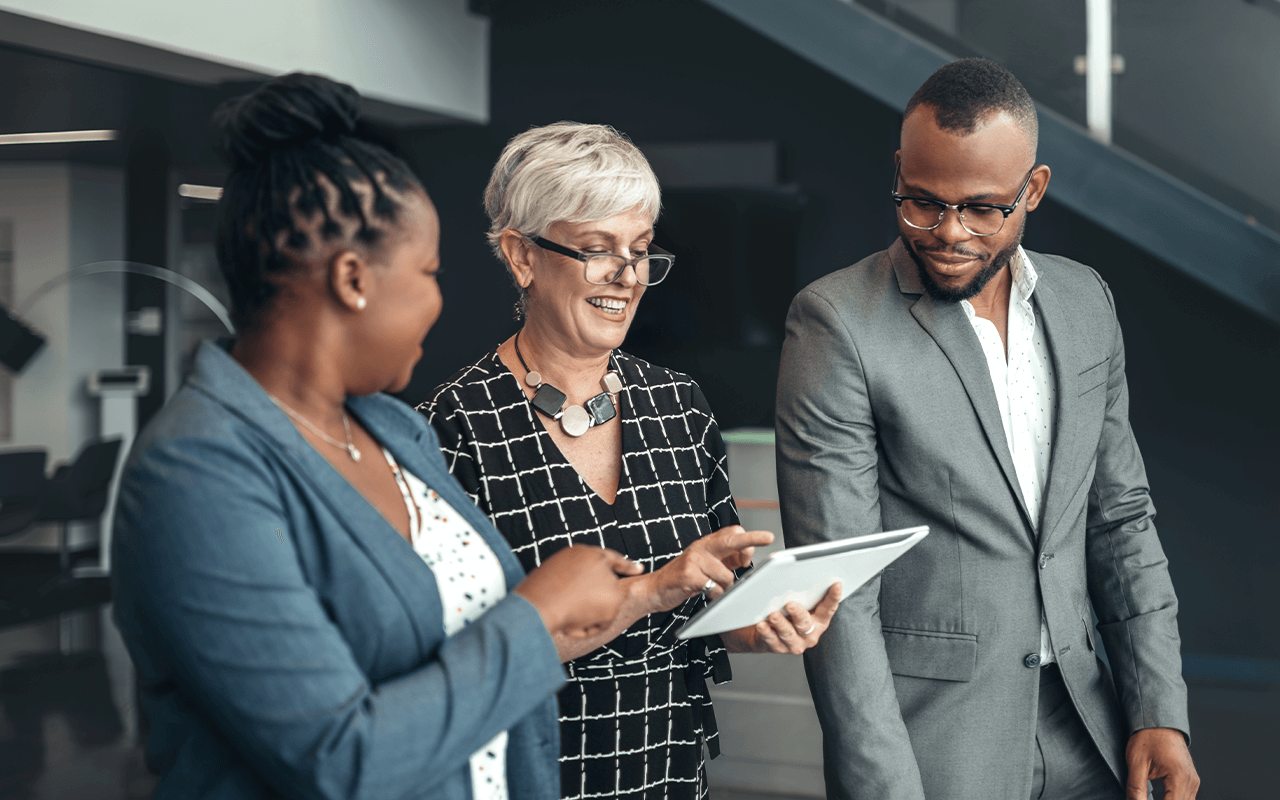 Three professionals in business attire discuss content on a tablet, standing in a modern office setting.