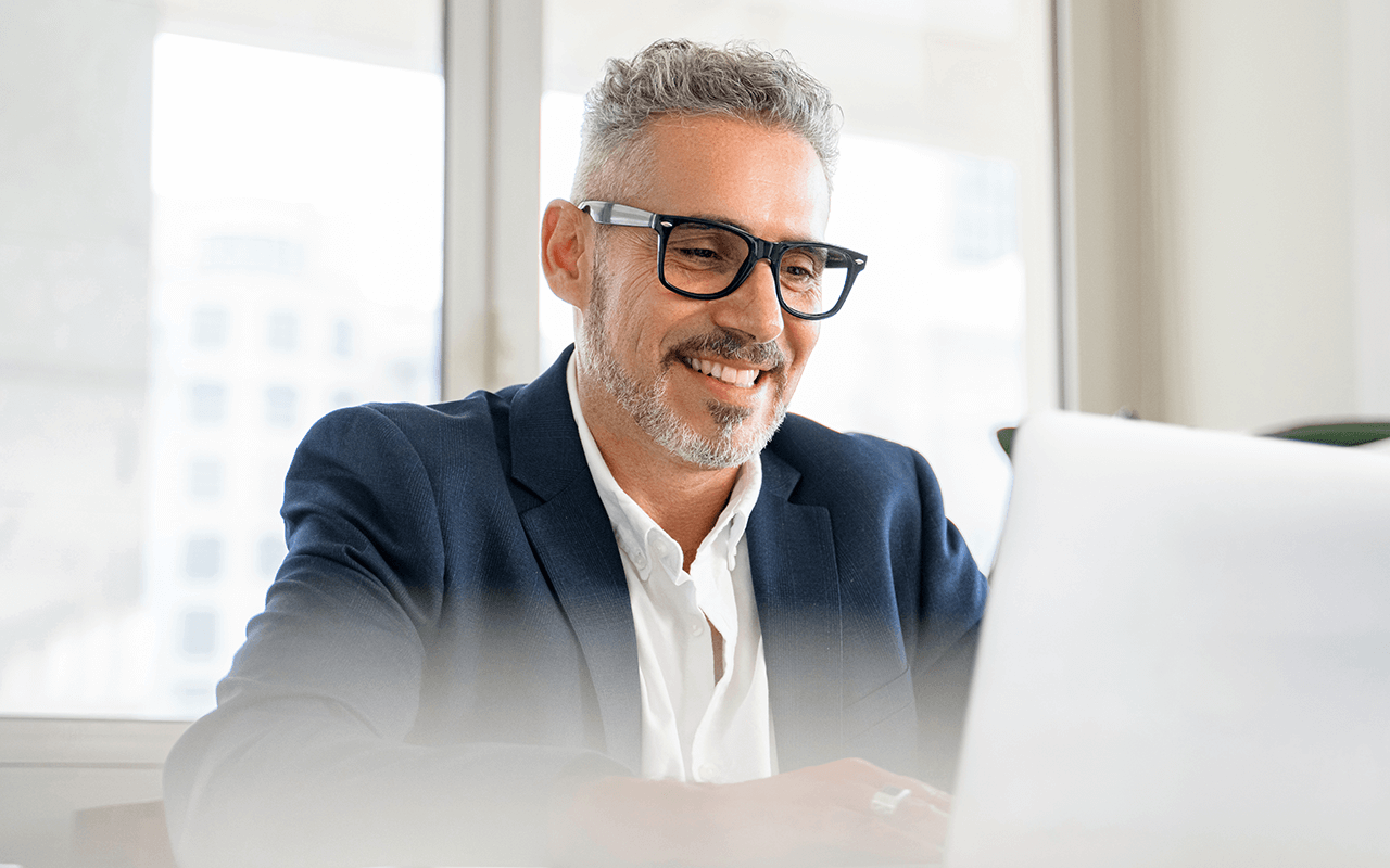 Smiling man in glasses and suit working on a laptop in a bright office setting.