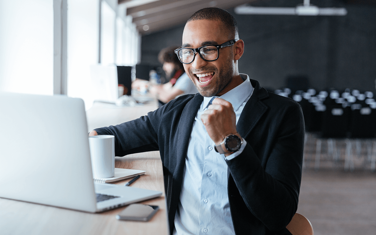A man in glasses and a suit smiles and raises his fist in excitement while looking at a laptop in a bright, modern office.