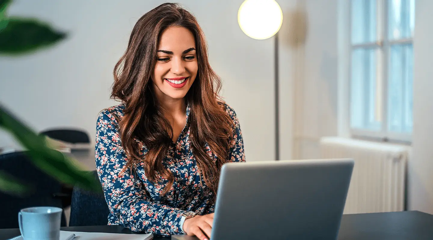Woman smiling while using a laptop at a desk with a cup and notebook, in a well-lit room with a standing lamp and window in the background.