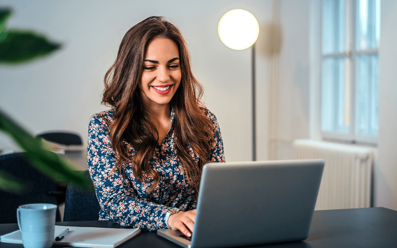 Woman smiling while using a laptop at a desk with a cup and notebook, in a well-lit room with a standing lamp and window in the background.