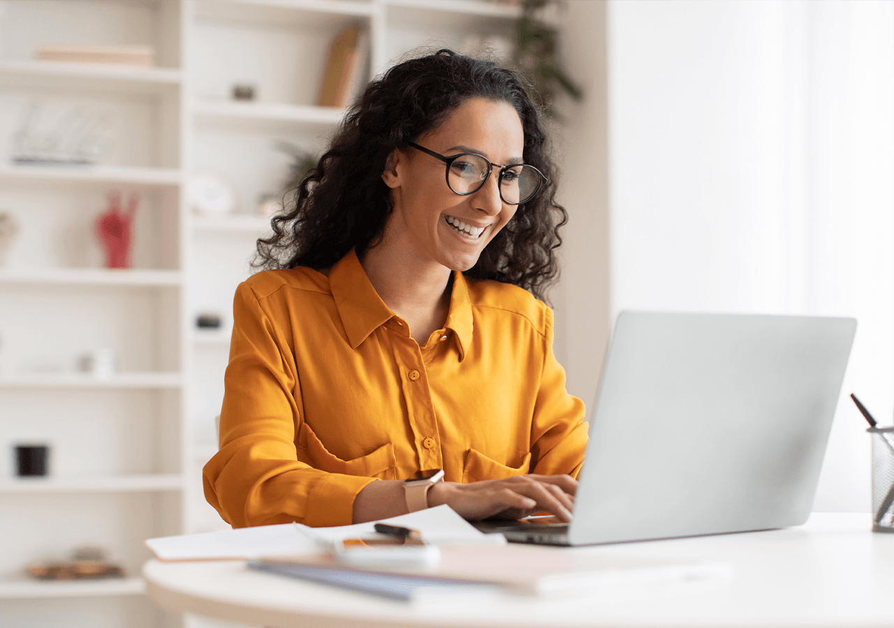 Smiling woman in a yellow shirt working on a laptop at a white desk, with shelves in the background.