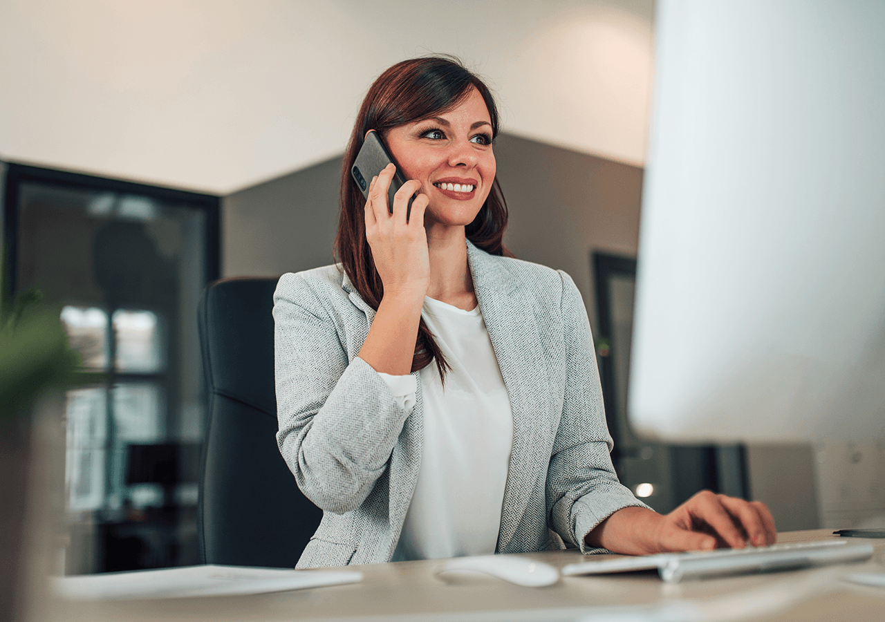 Smiling woman in a gray blazer talks on a phone while working at a computer in an office setting.