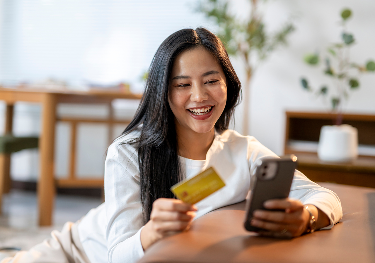 Woman smiling, holding a credit card and using her phone while lying on a couch in a cozy, well-lit room.