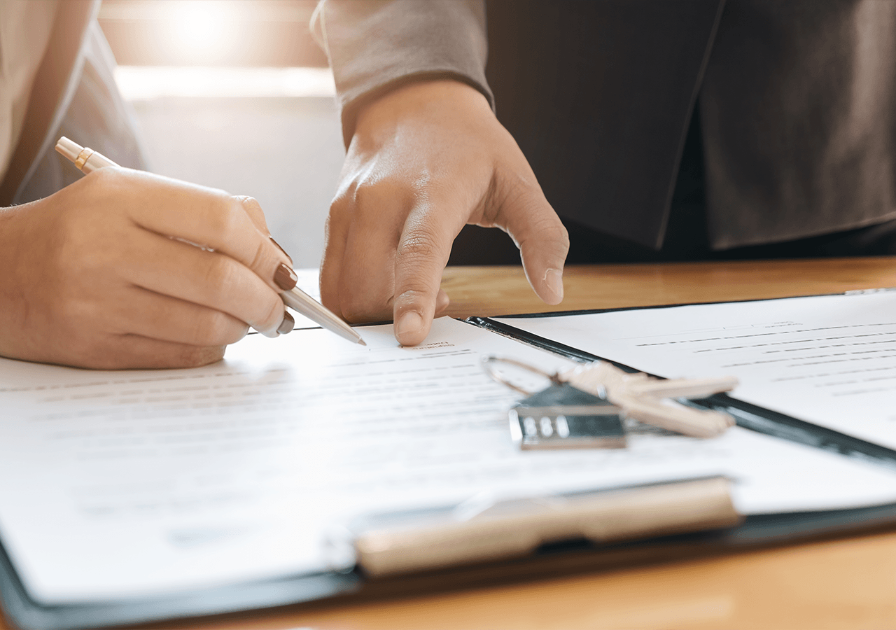 Person signing a document on a clipboard, guided by another person's pointing hand, with keys resting nearby.