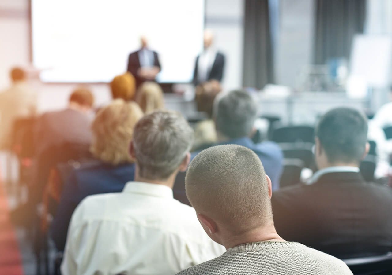 Audience listening to a presentation in a conference room. Two presenters stand at the front near a screen.