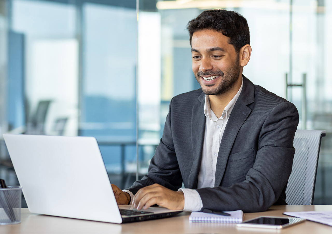 Smiling man in a suit working on a laptop at a desk in a modern office environment.