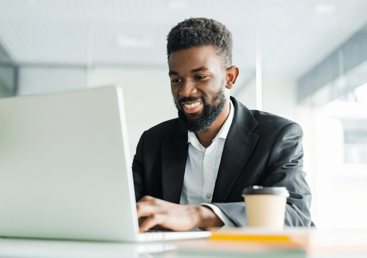 A man in a suit smiles while managing emails on a laptop in a law firm office, with a coffee cup nearby.
