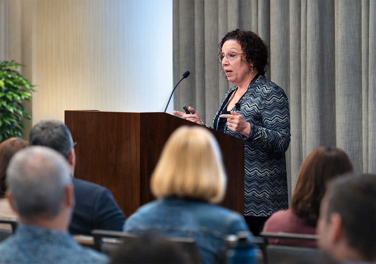 Christy A. Schmidt, Esq., speaking at a podium during a presentation, with an audience listening attentively.