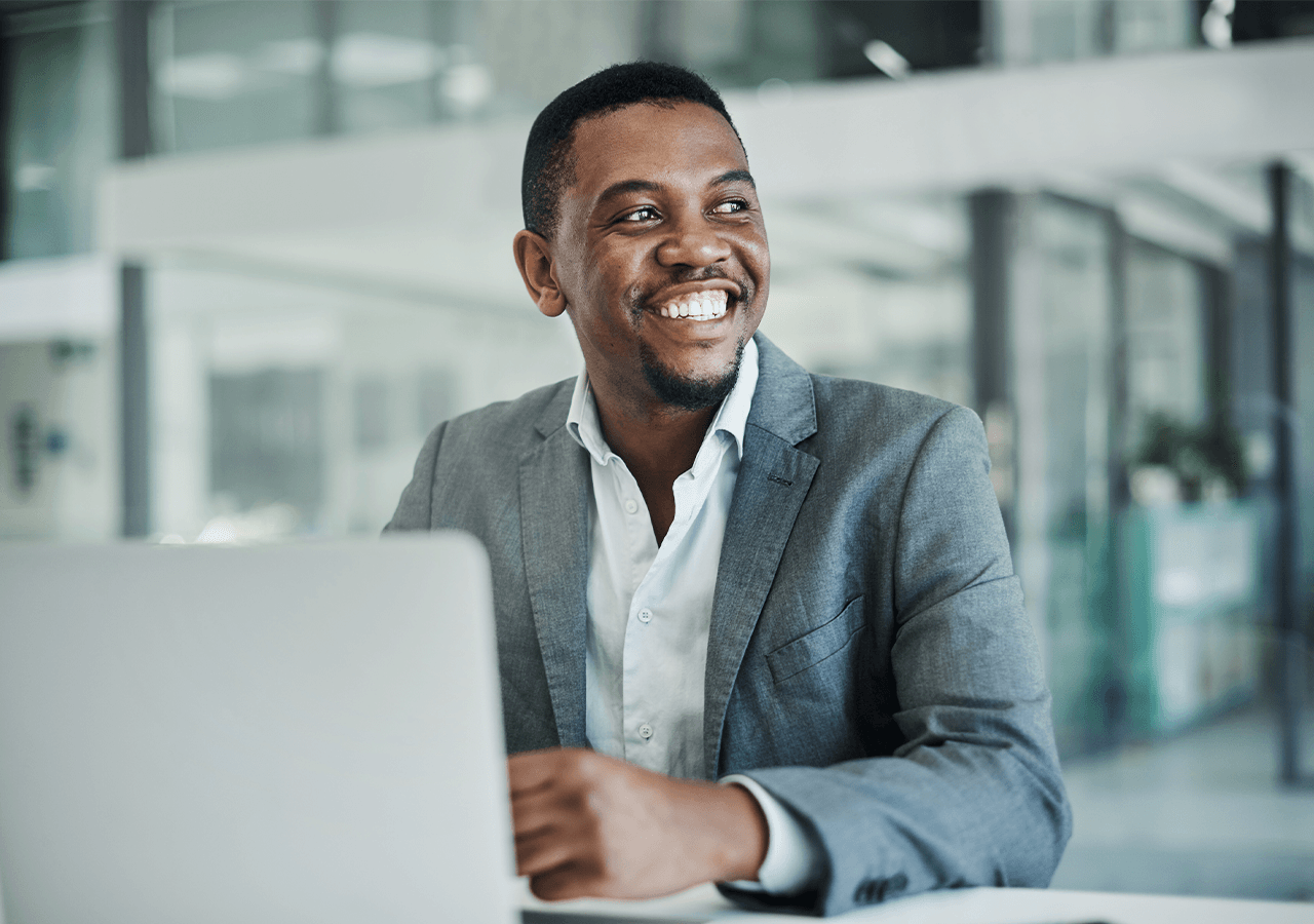 Smiling man in a gray suit sits at a table with a laptop, in a modern, bright office space.