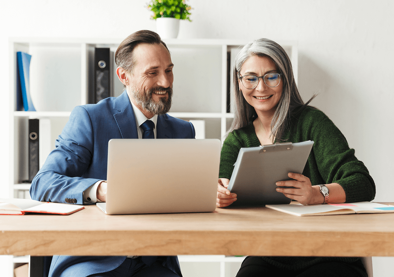 A man in a suit and a woman with glasses review documents on a clipboard at a desk with a laptop and books, smiling in a bright office.