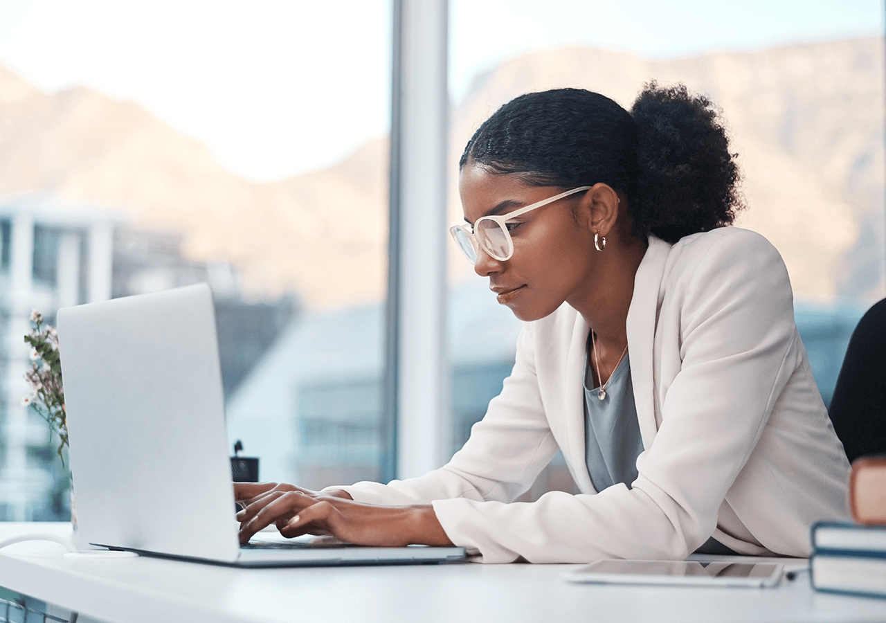A woman in a white blazer working on a laptop at a desk, with a window showing a mountainous view in the background.