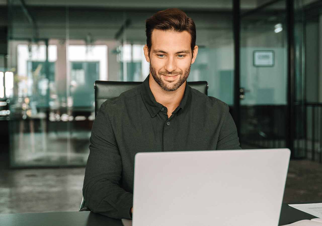 Man with a beard smiling while working on a laptop in an office setting with glass walls.