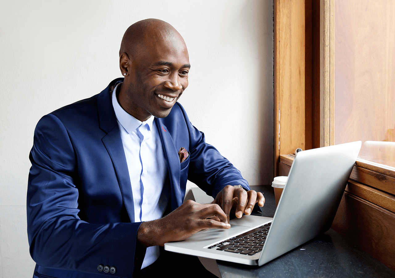 Smiling man in a blue suit working on a laptop near a window with a coffee cup beside him.