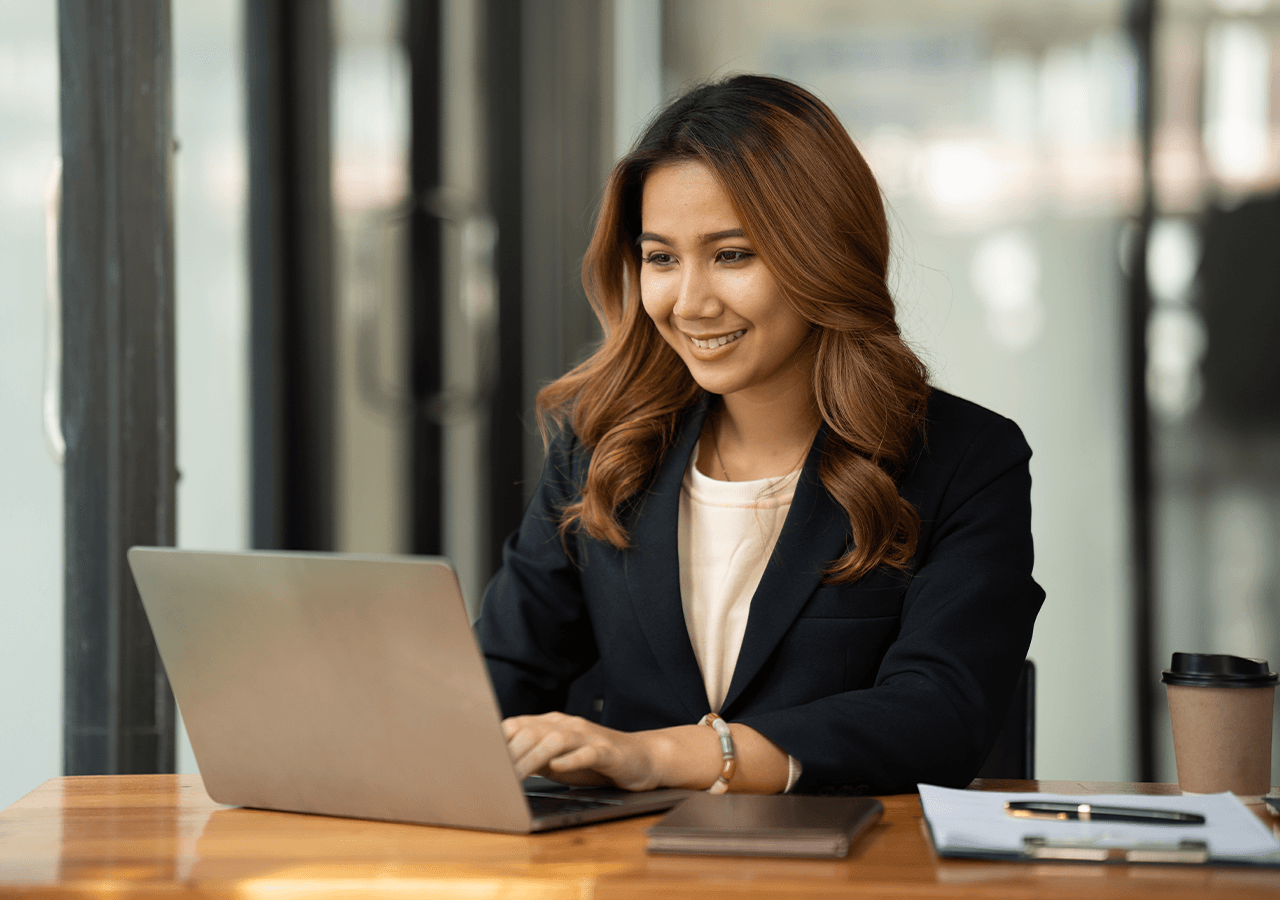 Smiling woman with long hair works on a laptop in an office setting, wearing a dark blazer. A coffee cup and notebook are on the wooden desk.