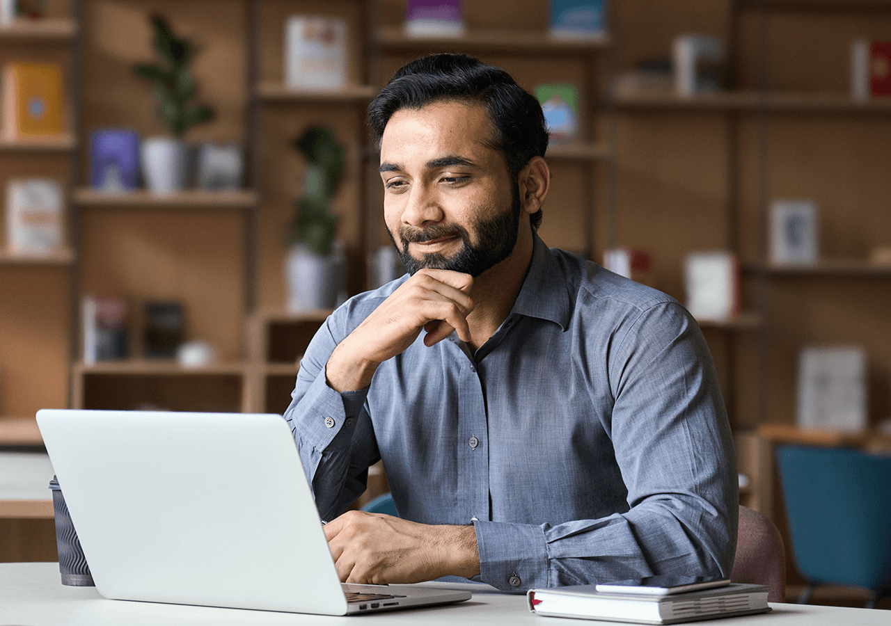 Smiling man in a blue shirt using a laptop at a desk, with bookshelves in the background.