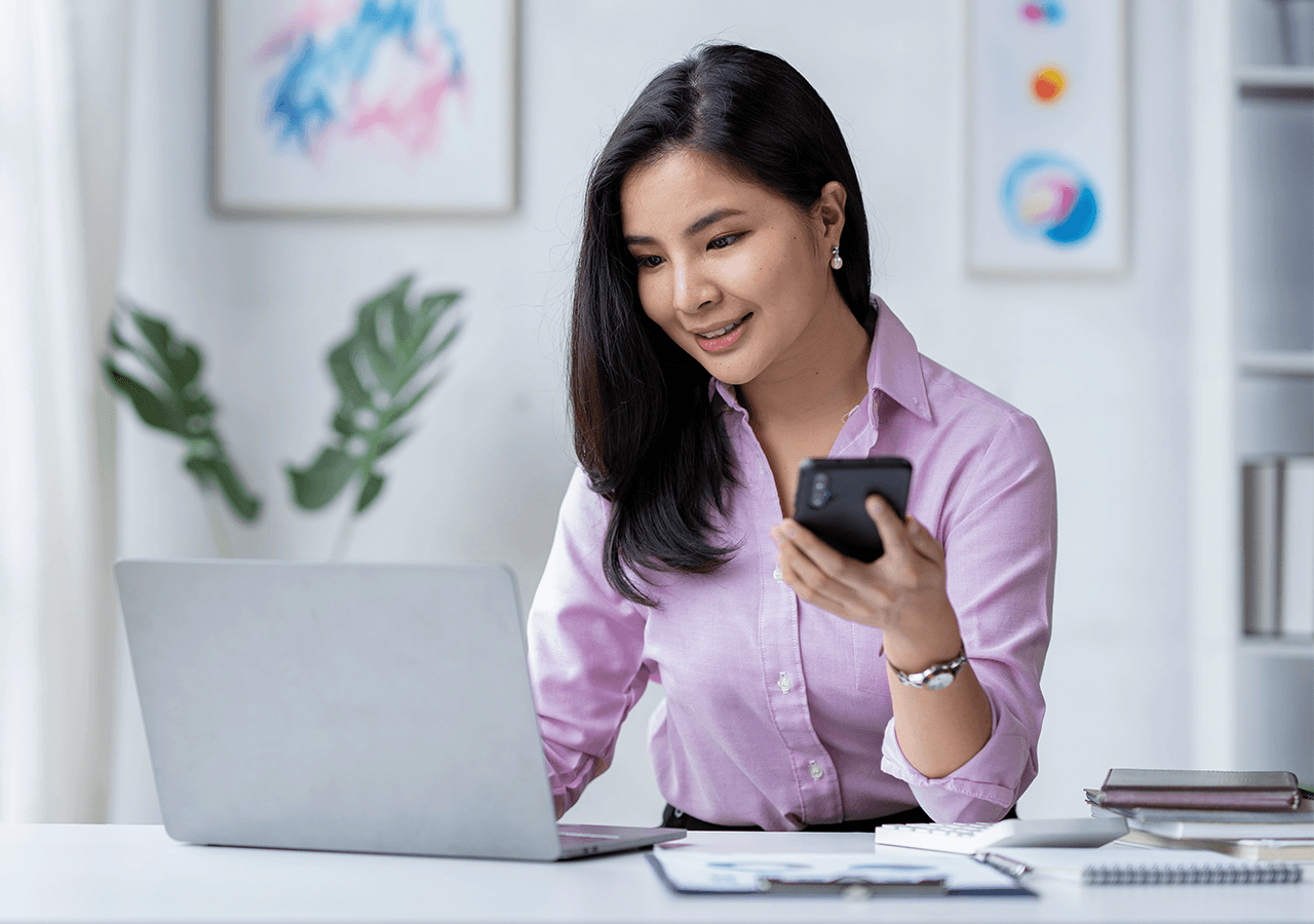 Woman in a pink shirt smiling while multitasking on a laptop and phone at a bright, modern office desk.