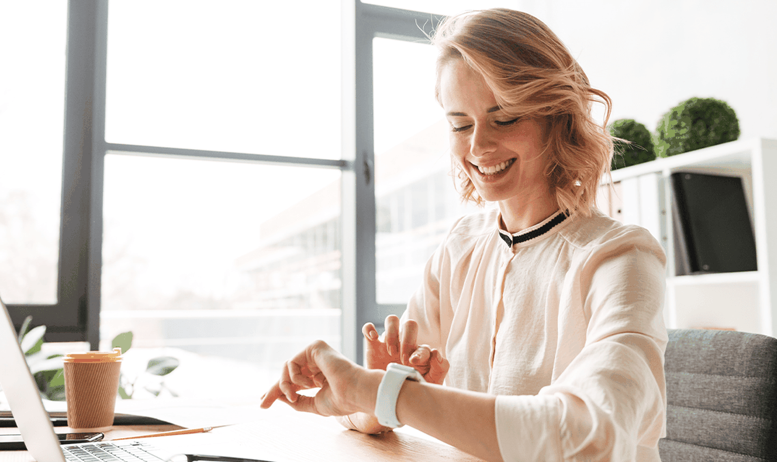 Smiling woman checks her smartwatch at a desk in a bright office, with a laptop, coffee cup, and window view.