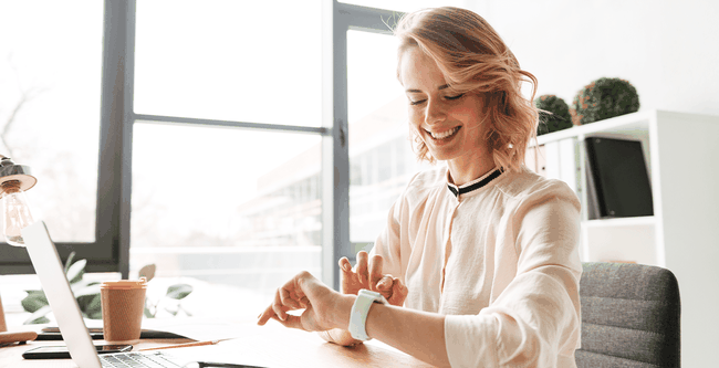 Smiling woman checks her smartwatch at a desk in a bright office, with a laptop, coffee cup, and window view.