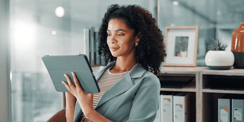 Woman with curly hair and a striped shirt smiles while holding a tablet, seated in a modern office with shelves and framed pictures behind her.
