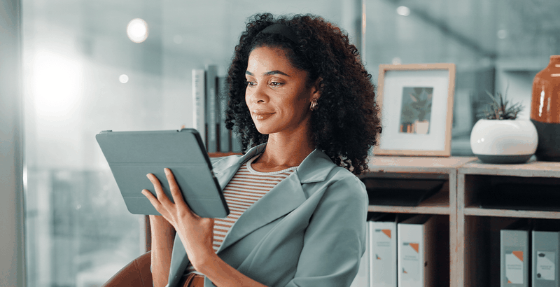 Woman with curly hair and a striped shirt smiles while holding a tablet, seated in a modern office with shelves and framed pictures behind her.