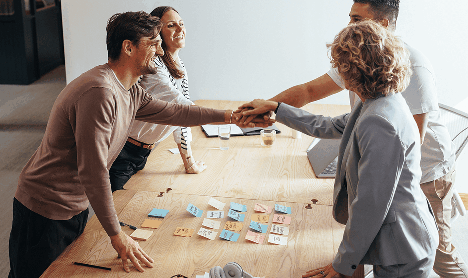 Four people in a meeting room, shaking hands around a table with sticky notes, notebooks, and headphones.