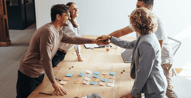Four people in a meeting room, shaking hands around a table with sticky notes, notebooks, and headphones.