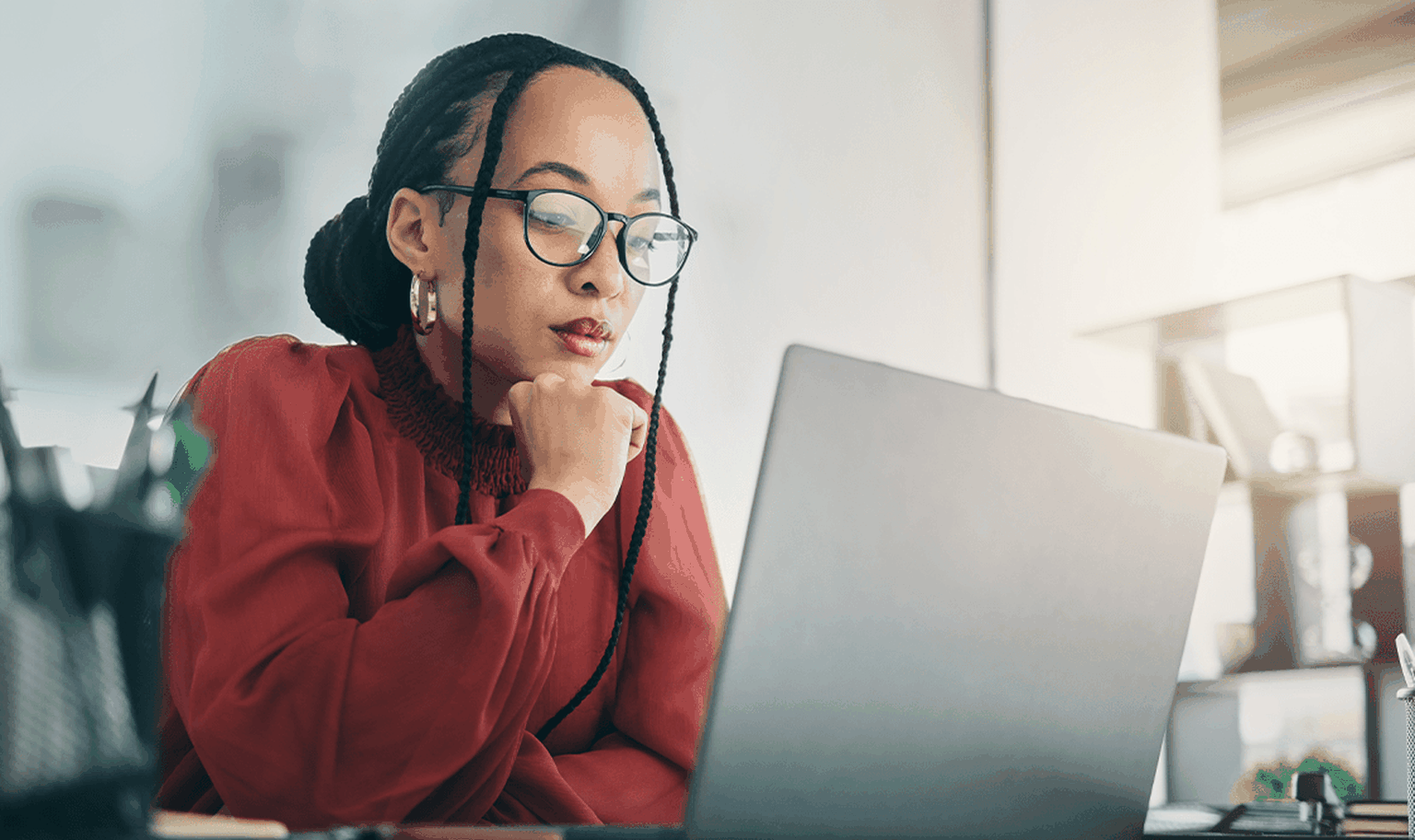 Woman in glasses and red blouse, focused on a laptop at a desk, with sunlight streaming through a window.