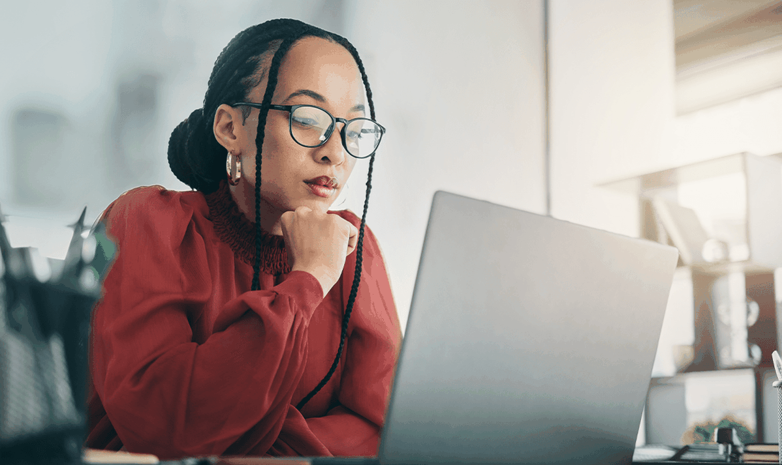 Woman in glasses and red blouse, focused on a laptop at a desk, with sunlight streaming through a window.