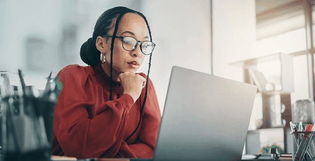 Woman in glasses and red blouse, focused on a laptop at a desk, with sunlight streaming through a window.