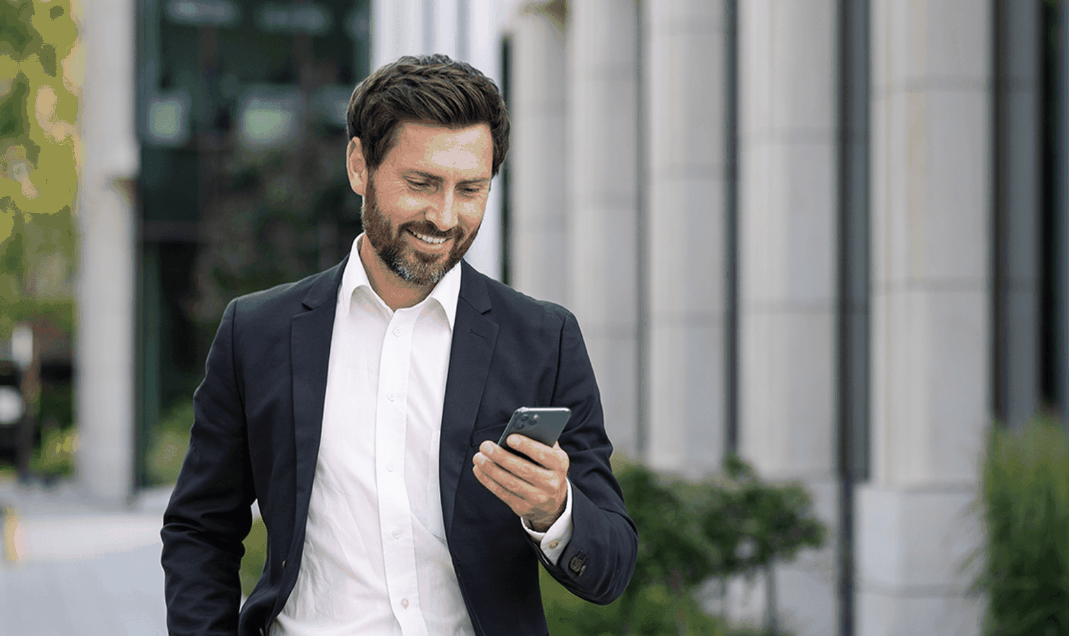 Smiling man in a suit holding a smartphone, walking outdoors near modern buildings and greenery.