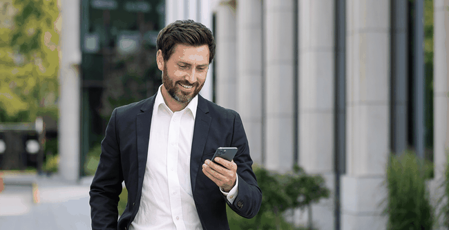 Smiling man in a suit holding a smartphone, walking outdoors near modern buildings and greenery.