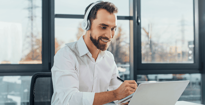 Man wearing headphones, smiling while working on a laptop in a bright office setting.