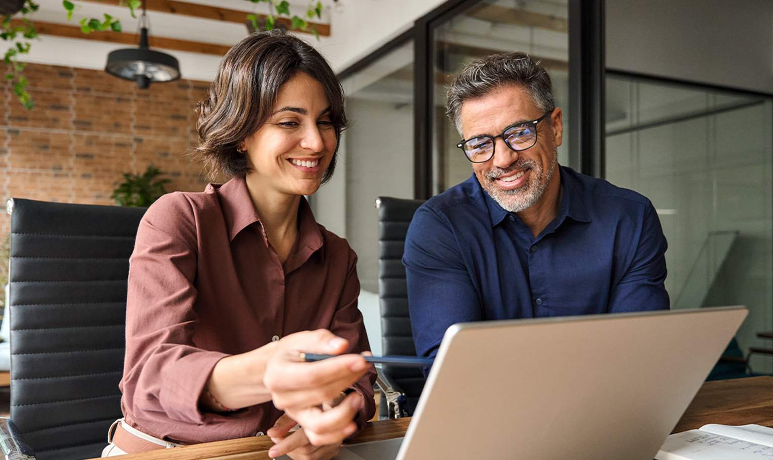 Two people smiling while looking at a laptop in a modern office setting with plants and brick wall background.