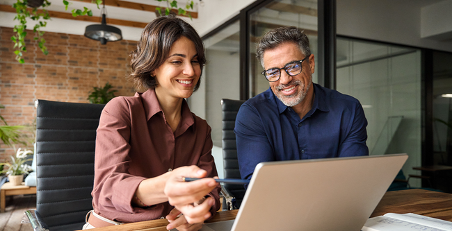 Two people smiling while looking at a laptop in a modern office setting with plants and brick wall background.