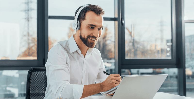 Man in white shirt wearing headphones, smiling while taking notes on a tablet at a desk with large windows in the background.