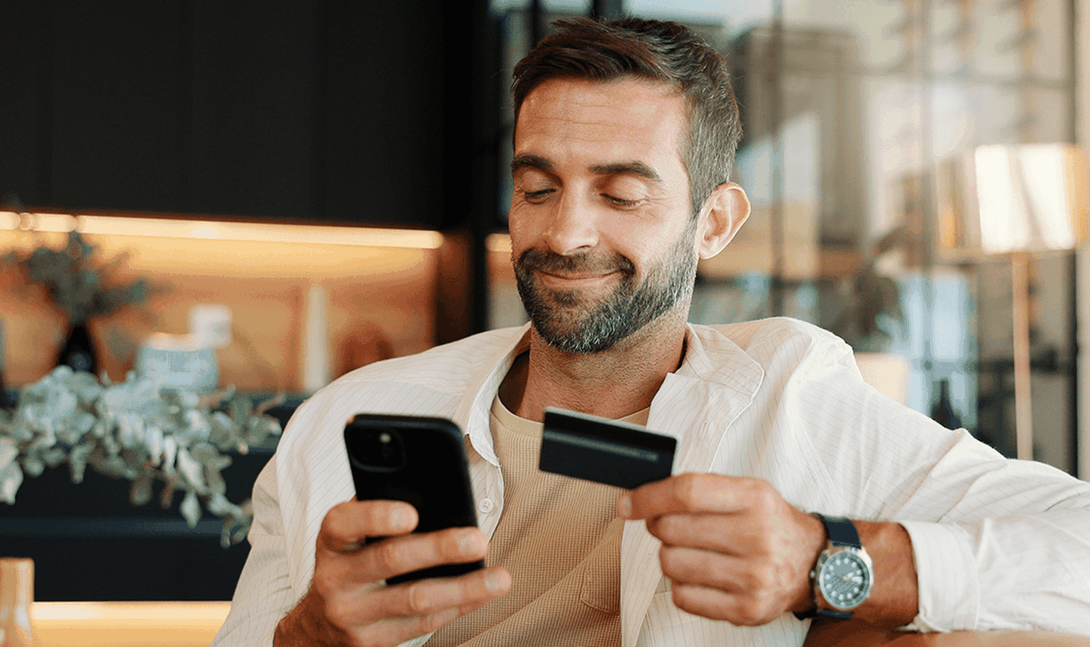Man smiling while using a smartphone and holding a credit card, seated in a modern living room.