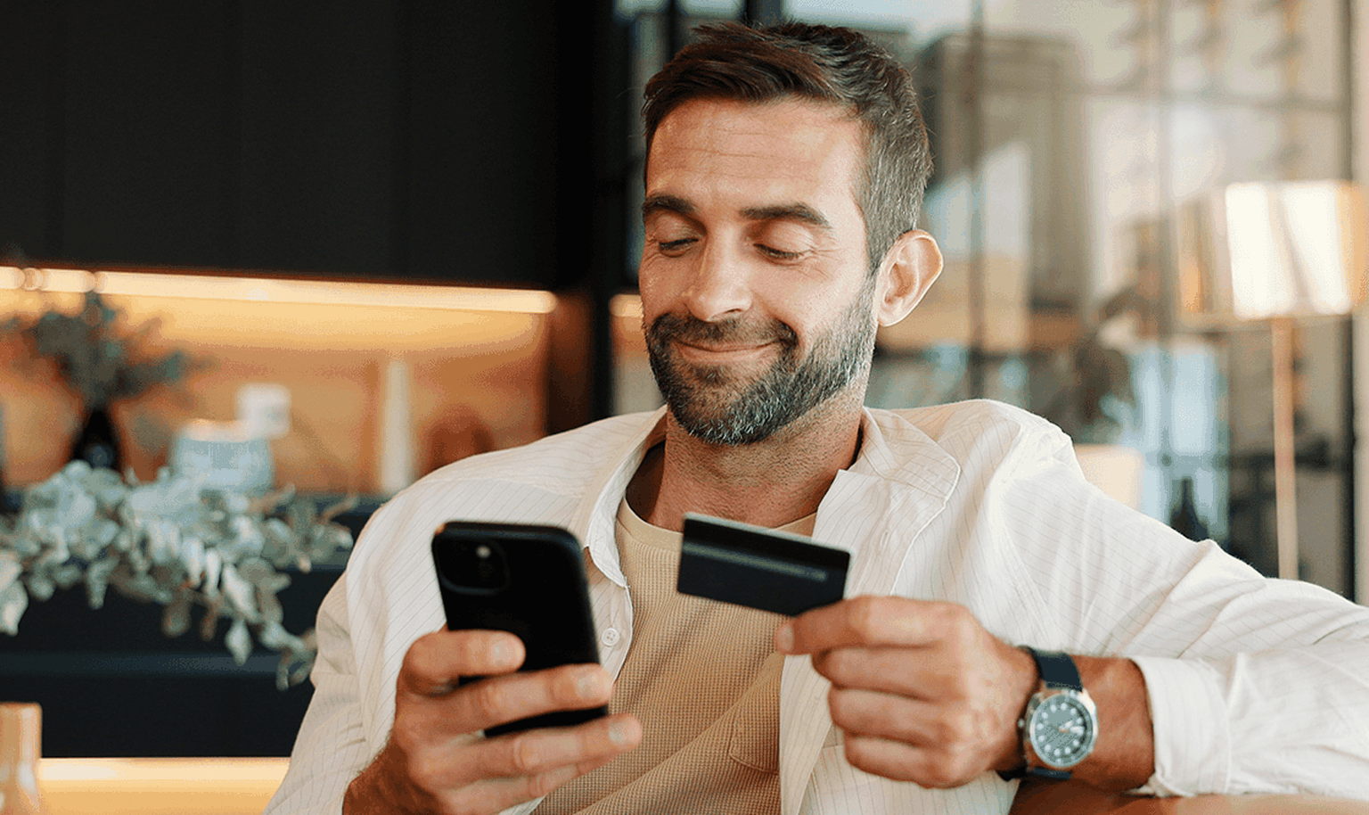 Man smiling while using a smartphone and holding a credit card, seated in a modern living room.