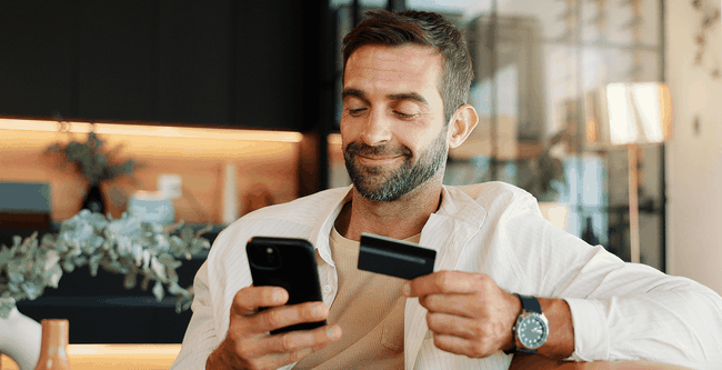 Man smiling while using a smartphone and holding a credit card, seated in a modern living room.