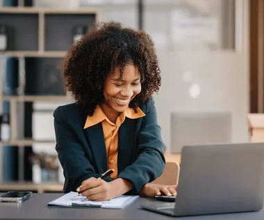 Smiling woman with curly hair works at a desk, writing on paper with a pen, laptop open in front, smartphone beside, office background.