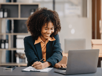 Smiling woman with curly hair works at a desk, writing on paper with a pen, laptop open in front, smartphone beside, office background.