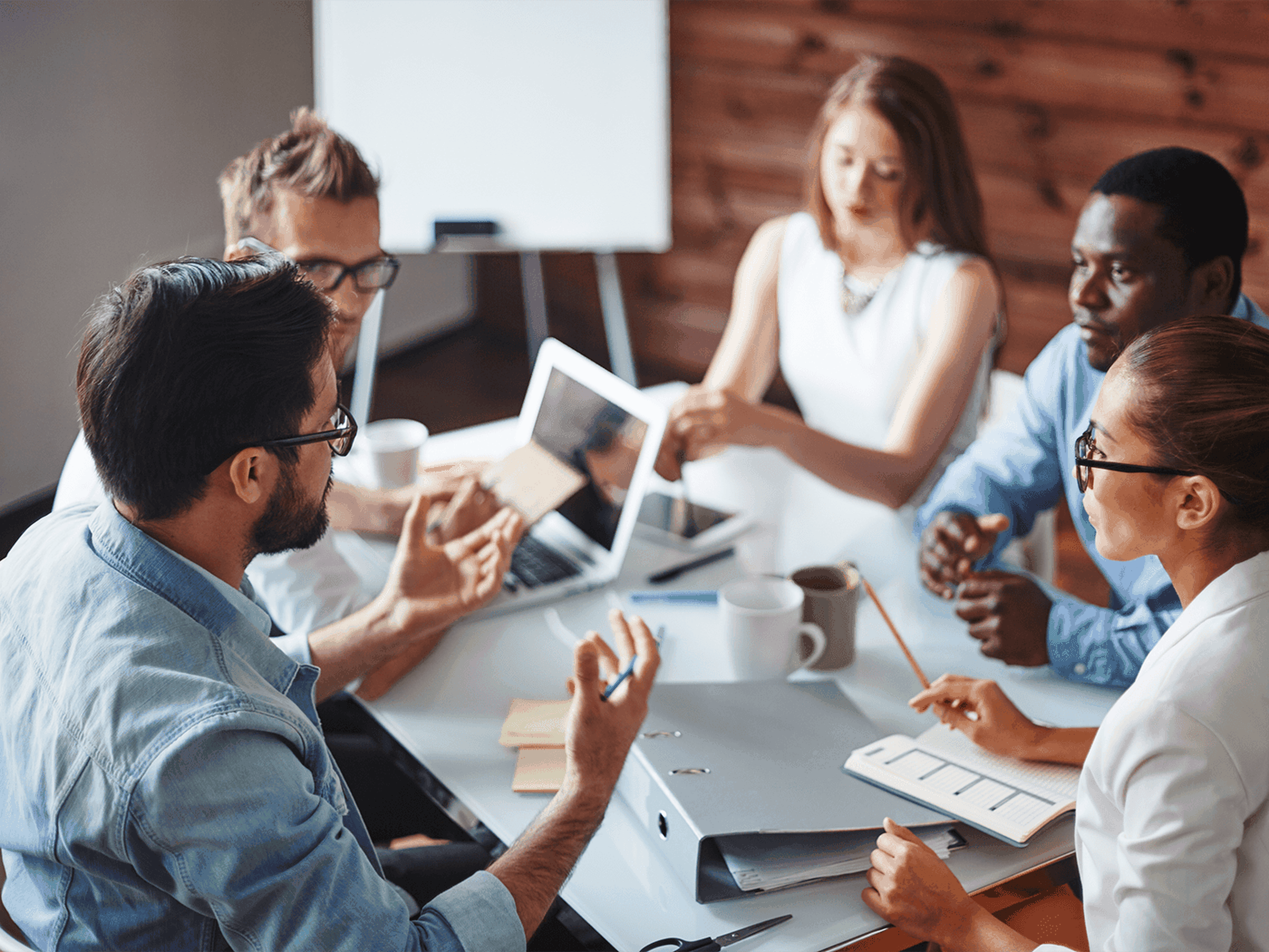 A group of five people having a discussion at a table with laptops, documents, and mugs. They are engaged in a collaborative meeting.