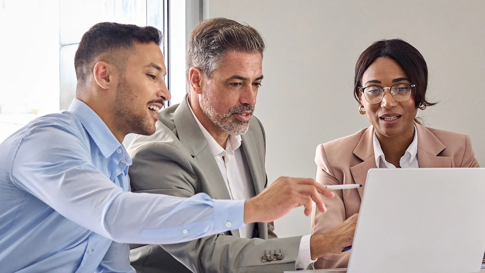 Three people in business attire are sitting at a table, engaged in a discussion, while looking at a laptop screen.