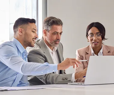 Three people in business attire are sitting at a table, engaged in a discussion, while looking at a laptop screen.