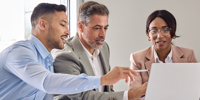 Three people in business attire are sitting at a table, engaged in a discussion, while looking at a laptop screen.