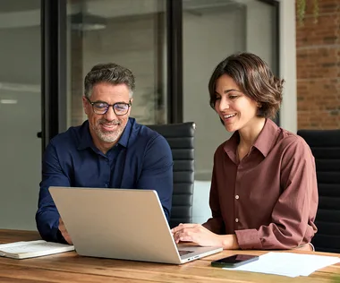 A man and a woman smiling and working together on a laptop at a wooden table in a modern office setting.